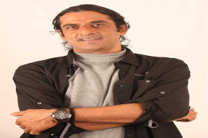 Ein Mann in einem schwarzen und aschfarbenen Kleid mit verschr├Ąnkten Armen, der eine Uhr tr├Ągt und gegen einen wei├čen Hintergrund steht, der nachdenklich erscheint.