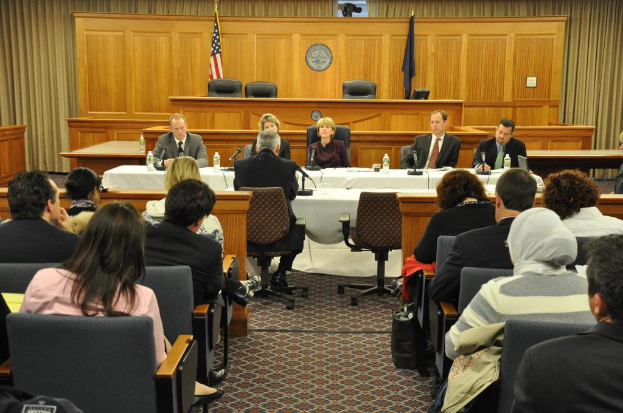 Ein Versammlungsraum mit einer Gruppe von Menschen, die sitzen, zwei ausländische Flaggen, ein oranger Schreibtisch mit Wasserflaschen und einem Mikrofon.