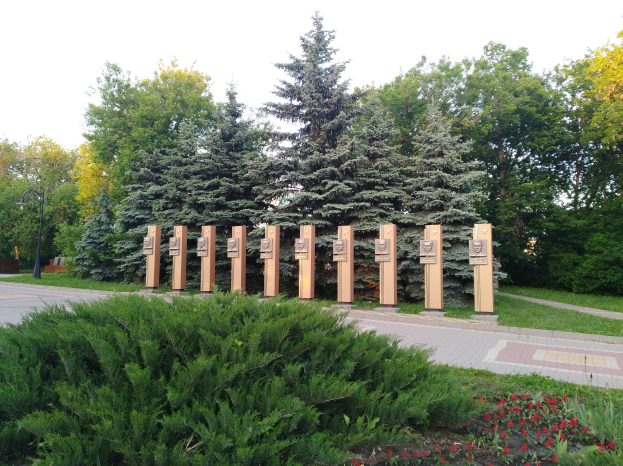 Ein Park mit einer Reihe von Bäumen in der Mitte, umgeben von blühenden Pflanzen, Gras, einem Weg, einem Straßenmast, einer Gedenktafel für die Opfer des Tschernobyl-Unfalls, einer Gruppe von Bäumen und einem bewölkten Himmel.