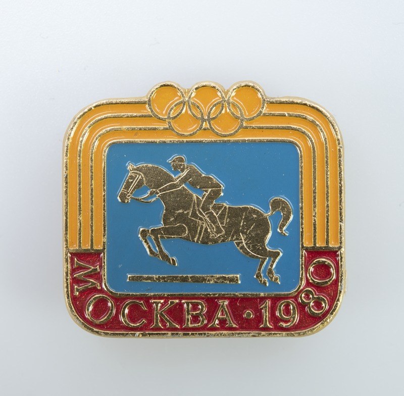 Olympische Spiele-Badge aus den 1988er Winterspielen mit einer Person in weißem Hemd und blauen Shorts auf einem rotbraunen Pferd mit weißer Mähne und Schweif gegen einem hellblauen Himmel mit weißen Wolken.