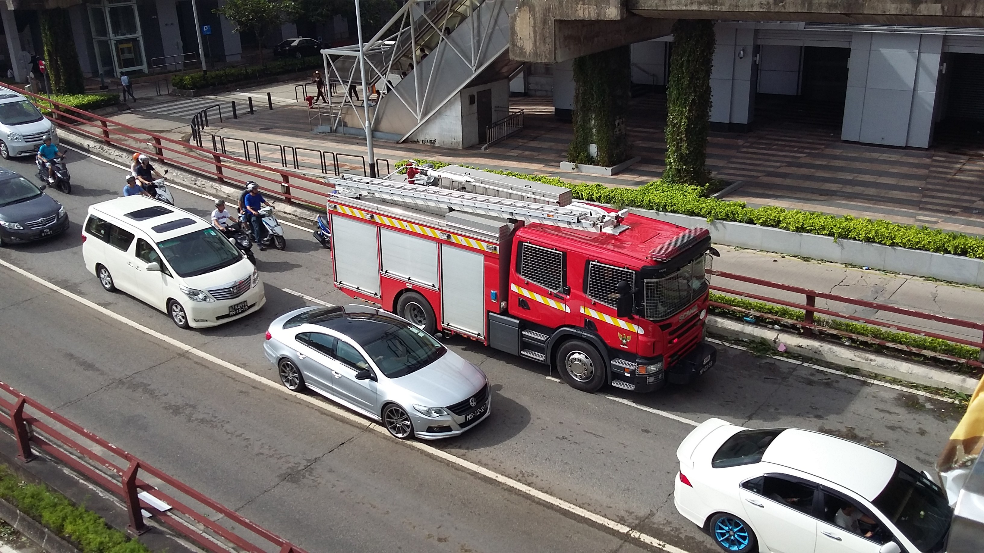 Feuerwehrfahrzeug fährt auf einer Straße mit einem Auto, Motorrädern und anderen Fahrzeugen; Gebäude, Bäume und Geländer im Hintergrund.