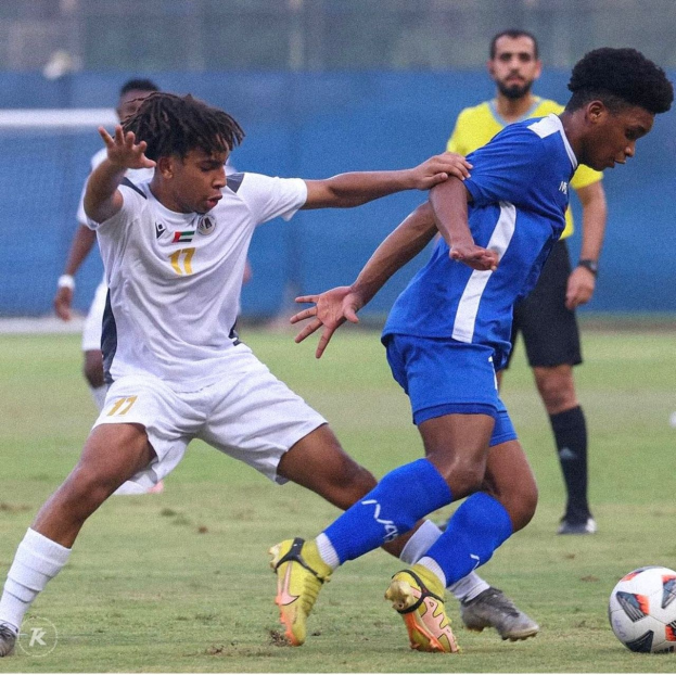 Zwei Männer beim Fußballspielen auf einem Rasenfeld mit einem Zaun im Hintergrund, einer scheint einen Sieg zu feiern.