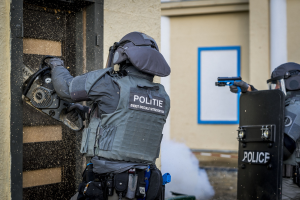 Zwei Polizisten in Einsatzausrüstung stehen vor einem Gebäude, einer hält eine Waffe und der andere ein Objekt, mit einer Tür links und einer Tafel an der Wand im Hintergrund.