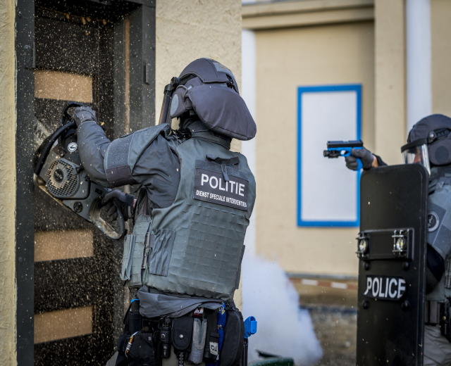 Zwei Polizisten in Einsatzausrüstung stehen vor einem Gebäude, einer hält eine Waffe und der andere ein Objekt, mit einer Tür links und einer Tafel an der Wand im Hintergrund.