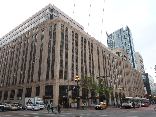 Eine Straße mit Menschen, die Fahrräder, Busse und Autos fahren; im Hintergrund gibt es Bäume, Gebäude, Lichter, Pfosten, Drähte und den Himmel.