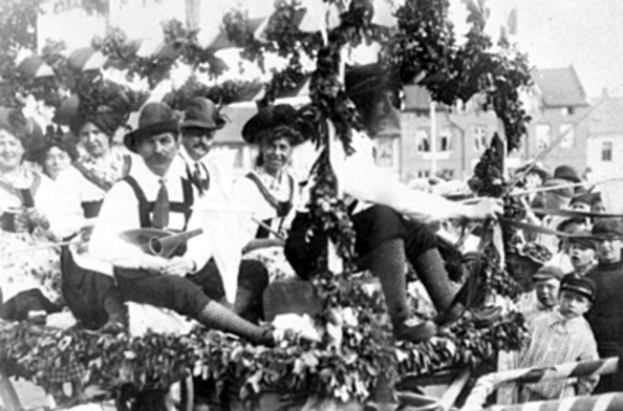 Eine Gruppe von Menschen in traditioneller bayrischer Tracht fährt auf einem mit Blättern geschmückten Festwagen in einem Umzug mit, einige spielen Musikinstrumente, mit Gebäuden und einem klaren Himmel im Hintergrund, dargestellt in Schwarz-Weiß.