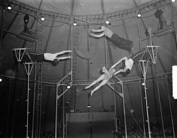 Schwarzes und weißes Foto von Akrobaten, die in einem Zirkuszelt performen, während das Publikum unten zusieht.