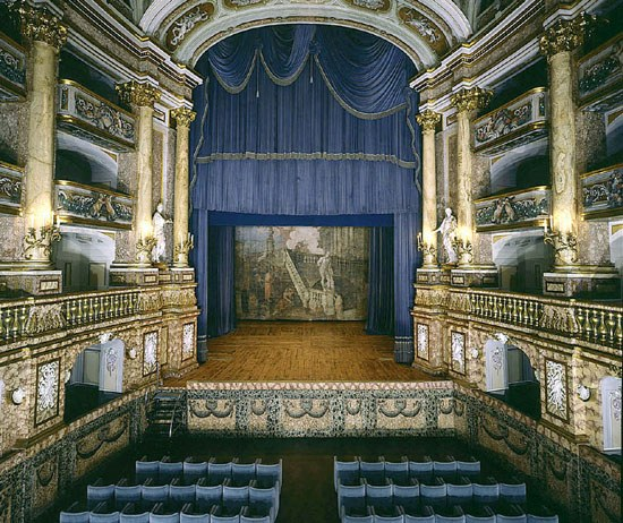 Auditorium mit blauen Stühlen, einem blauen Vorhang, kunstvoll gestalteten Wänden mit Skulpturen, geschnitzter Decke und hell erleuchteter Bühne.