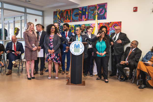 Eine Gruppe von Menschen, die vor einem Rednerpult mit Mikrofon und einer Tafel mit der Aufschrift "Sancho-Stadtrat tritt dem Stadtrat bei" stehen und sitzen, mit Flaggen, einer Tafel, einem Feuerlöscher und Deckenleuchten an der Rückwand.