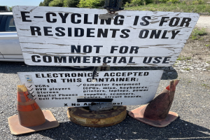 Schild mit Aufschrift "E-Bike-Fahren nur für Anwohner" neben einer Straße mit Verkehrskegeln, einem Fahrzeug, Bäumen und einem klaren blauen Himmel im Hintergrund.