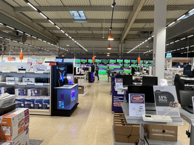 Ein großer Elektronikbereich in einem Walmart-Laden, mit Regalen voller Computer, Drucker, Kartons und anderen Geräten, sowie Menschen, die durch die Gänge schlendern und Deckenventilatoren zu sehen sind.
