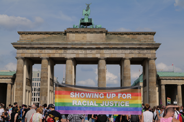 Eine Gruppe von Menschen steht vor dem Brandenburger Tor in Berlin, Deutschland, mit einer Tafel, auf der 'Rassengerechtigkeit' steht, im Hintergrund sind Säulen, eine Statue und Gebäude zu sehen.