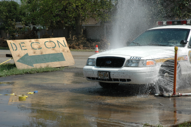 Ein Polizeiwagen fährt durch eine Pfütze, mit einem Schild, einem Verkehrskegel und anderen Gegenständen auf dem Boden, vor einem Hintergrund aus Bäumen, Gebäuden und einem klaren blauen Himmel.