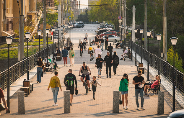 Eine Gruppe von Menschen, die ├╝ber einen Gehweg neben einem Zaun gehen und Fahrr├Ąder fahren, mit anderen, die auf B├Ąnken sitzen, Fahrzeugen auf der Stra├če, Laternenmasten, Strommasten mit Kabeln, Schildern, B├Ąumen, Geb├Ąuden mit Fenstern und einem klaren blauen Himmel im Hintergrund.