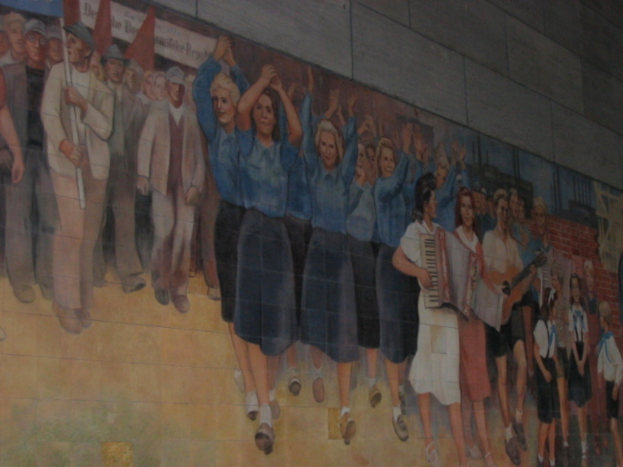 Eine Gruppe von Menschen mit blauen Mädchen in der Mitte, die klatschen, Musikern auf der rechten Seite, spielenden Kindern in der rechten Ecke und Personen mit roten Fahnen auf der linken Seite.