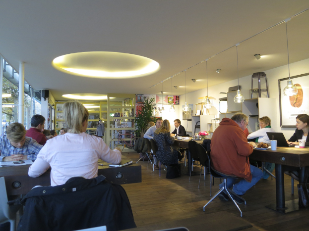 Ein Arbeitsplatz mit mehreren Tischen und Stühlen, Menschen, die darum arbeiten, ein Foto auf der rechten Wand, Schränke, hängende Lampen, eine Pflanze rechts, eine Schaufensterpuppe links und eine Tür im Hintergrund.