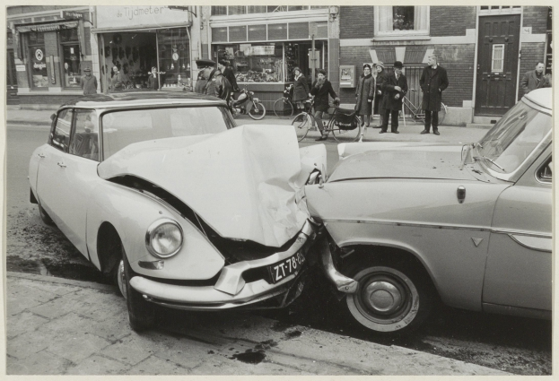 Schwarz-weiß-Foto von zwei beschädigten Autos am Straßenrand mit Menschen und Fahrrädern in der Nähe, Gebäude im Hintergrund sichtbar.
