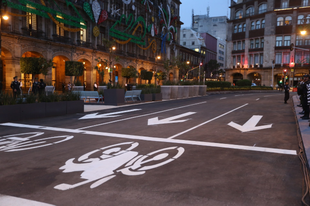 Eine Stadtstraße mit einem ausgewiesenen Fahrradweg, gesäumt von Gebäuden, Straßenmöblierung, Fußgängern auf dem Gehweg, Grünfläche und einem klaren blauen Himmel.