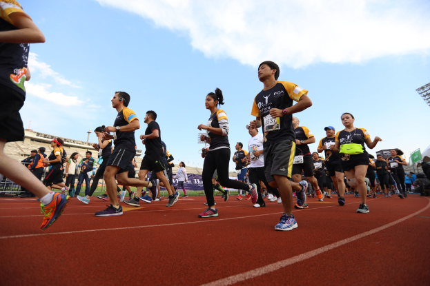 Gruppe von Menschen, die auf einer Laufbahn laufen, mit Zuschauern im Hintergrund, vor Gebäuden, Laternenmasten, Bannern und einem klaren blauen Himmel während eines Rennens.