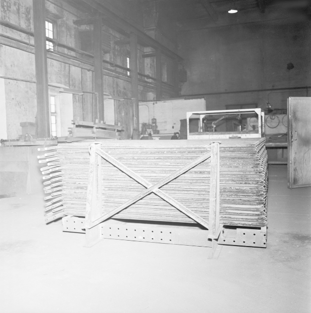 Ein Schwarz-Weiß-Foto einer Eisengießerei mit einem großen Metallgerüst, Metallobjekten auf dem Boden, einer Tür auf der rechten Seite, einer Wand mit Fenstern im Hintergrund und Lampen oben.