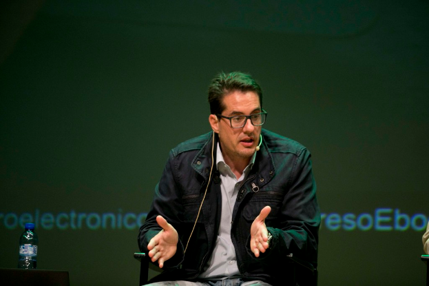 Eine Person sitzt in der Mitte auf einem Stuhl und hält eine Rede, während sie ein Mikrofon trägt, mit einer Flasche mit blauer Kappe auf einem Tisch links und einem Bildschirm im Hintergrund.