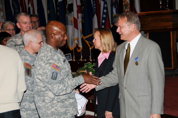 Eine Gruppe von Menschen mit zwei Personen, die sich die Hände schütteln, Fahnen im Hintergrund, ein Podium mit einem Mikrofon und eine Topfpflanze in der Nähe.