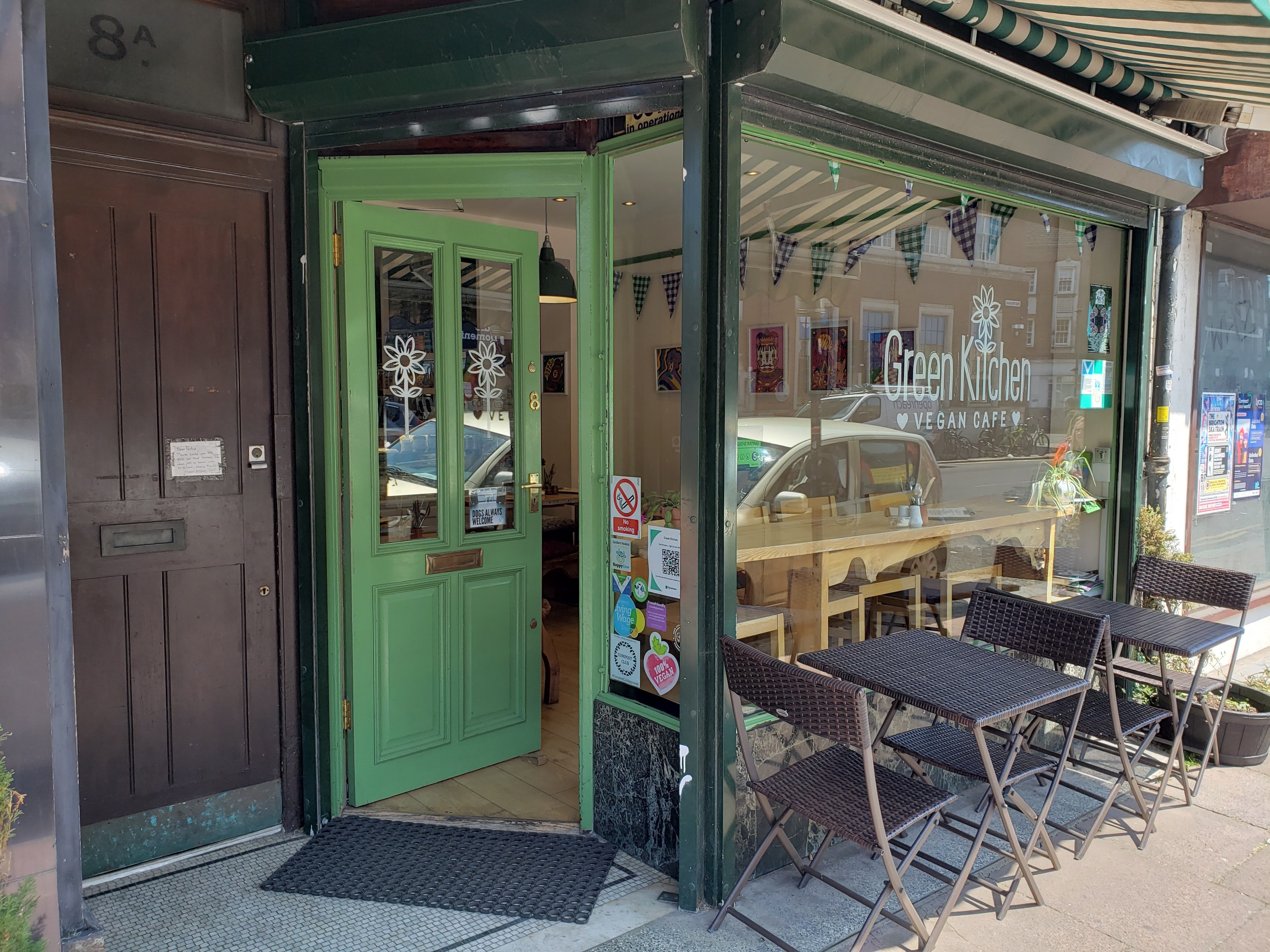 Außensitzbereich des Green Kitchen Vegan Cafe mit Tischen, Stühlen, Pflanzen, einer Fußmatte und einer Glastür mit Postern; drinnen sind Tische, Stühle und eine Deckenleuchte zu sehen.