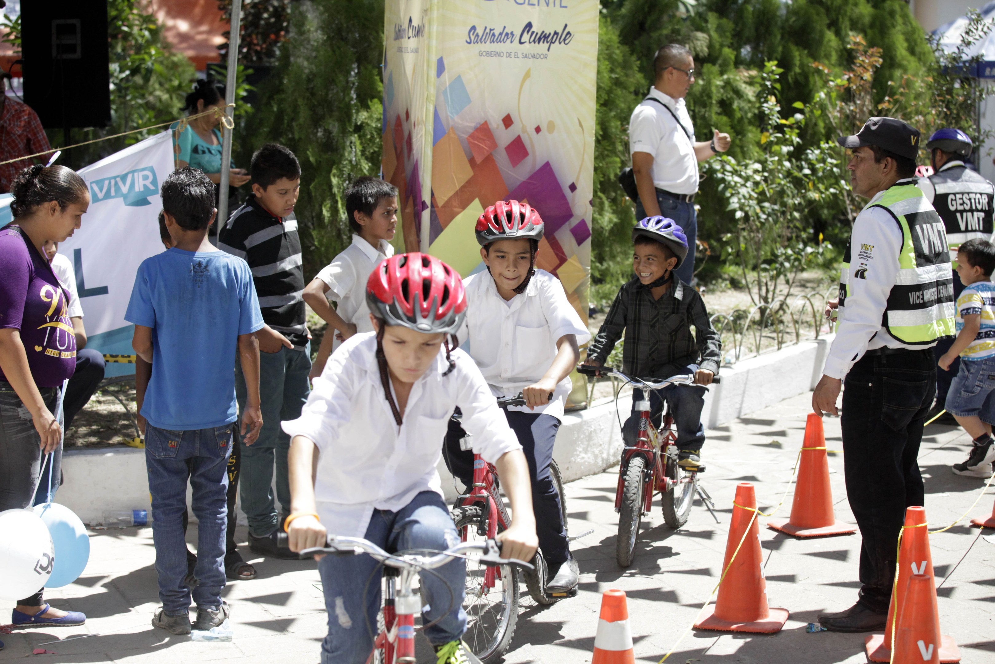 Eine Gruppe von Kindern fährt Fahrräder auf einer Straße mit Verkehrskegeln, einige tragen Helme, andere stehen in der Nähe, mit einer Fahne, Bäumen und Gebäuden im Hintergrund.