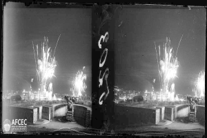 Ein Schwarz-Weiß-Foto von Feuerwerk, das über einer nächtlichen Stadt explodiert, während des Catalunya-Feuerwerksfestivals in Mexiko-Stadt, Mexiko im Jahr 1939, mit Menschen und Gebäuden im Hintergrund.