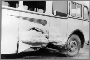 Schwarzes und weißes Foto eines verunglückten Busses mit eingedellter Front, zersplitterten Fenstern und geöffneter Fahrertür, die das Innere zeigt.