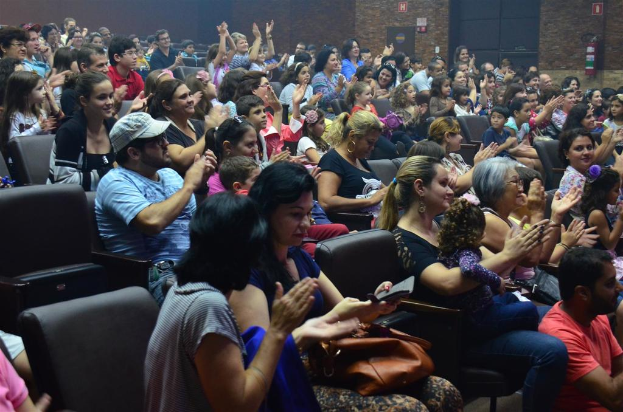 Eine Gruppe von Menschen, die auf Stühlen sitzen, lächeln, einige halten Gegenstände, tragen Brillen und einer trägt eine Mütze, während andere klatschen, mit einer Wand, Schildern und einem Feuerlöscher im Hintergrund.