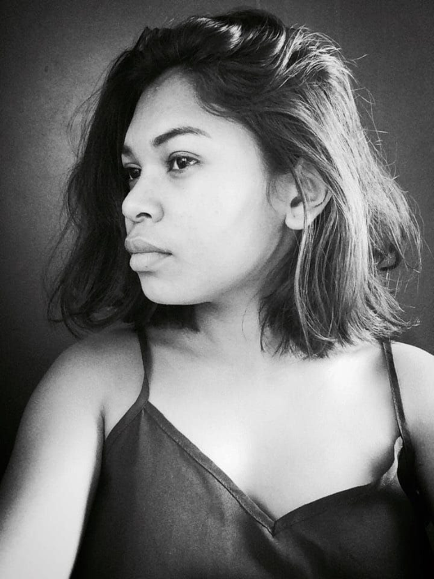 Eine selbstbewusste Frau in einem schwarzen Kleid posiert in einem Schwarz-Weiß-Foto vor einer Wand mit einem glatten Bob und dezentem Make-up.