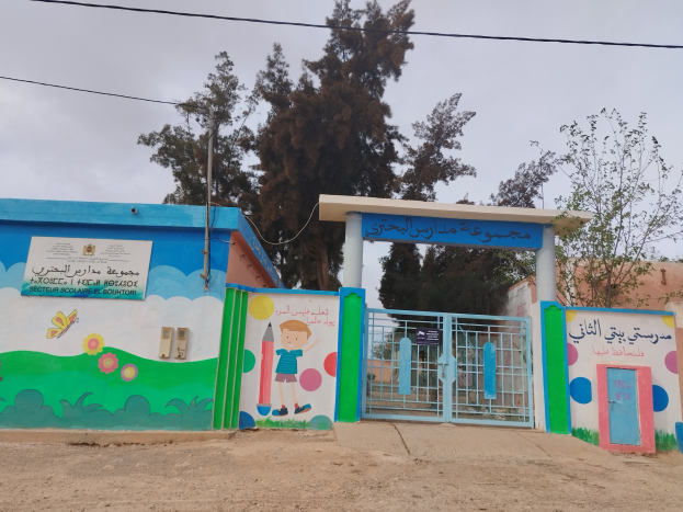 Blau-weißes Gebäude mit Wandgemälde, das als Eingang zu einer Kinder Schule dient, mit einem Tor, einem Namensschild, umgeben von Häusern, Bäumen, einem Strommast mit Drähten und einem bewölkten Himmel.