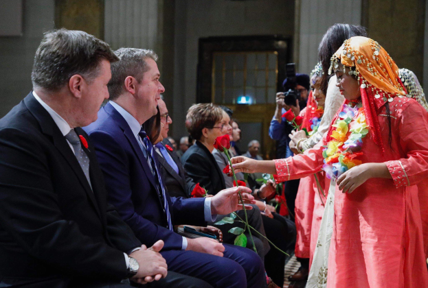 Eine Gruppe von Menschen sitzt zusammen in einem Raum, mit einer Frau in der Mitte, die einen Kranz trägt und Blumen hält, flankiert von einem Mann in einem Anzug und jemandem, der eine Kamera hält, vor einer Wand mit einer Tür.