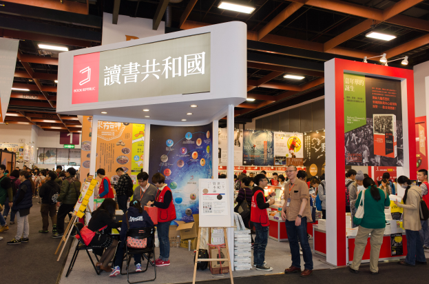 Eine große Gruppe von Menschen an einem Messestand mit Schildern und Tafeln, die "Book Republic Hong Kong Book Fair" lesen, einige sitzen, einige stehen, unter einem Dach mit Deckenbeleuchtung.