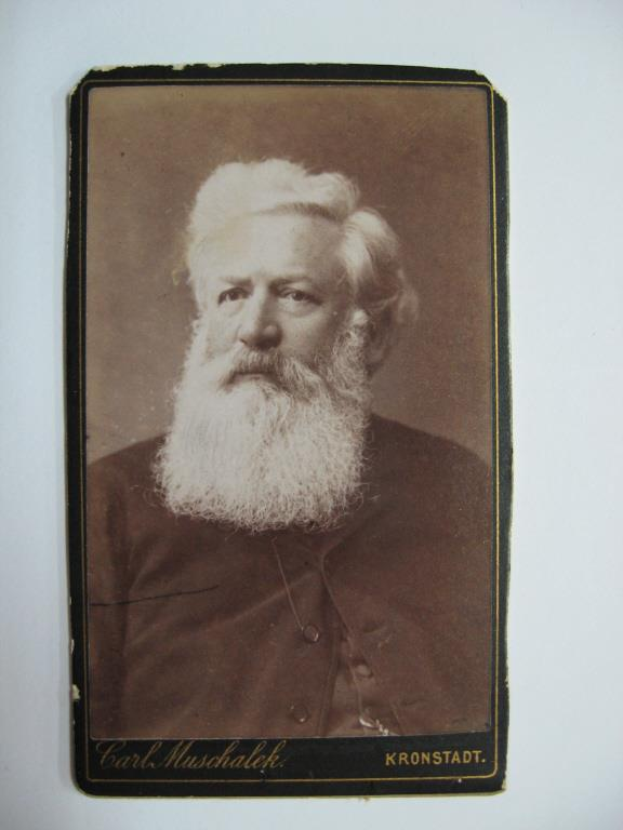 Schwarzes und weißes Foto von Carl Muschalk, einem deutschen Komponisten mit langem weißem Bart, befestigt an einer weißen Oberfläche mit Text unten.