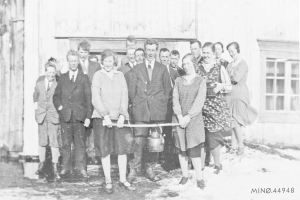 Schwarzes und weißes Foto von Menschen, die strahlend eine Schnur halten, vor einem Gebäude mit einem Fenster und einer Tür, wahrscheinlich bei der Einweihung einer neuen Schule.