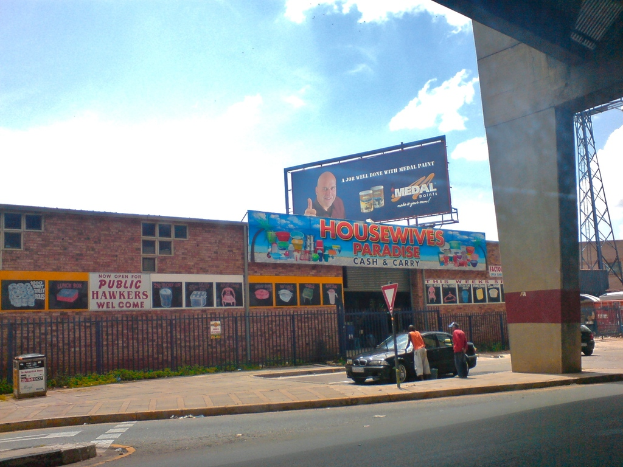 Eine Straße mit einer Säule und einem Auto auf der rechten Seite, ein Mast mit einer weißen und roten Tafel, ein Auto mit zwei Männern daneben, ein Müllcontainer auf der linken Seite, braune Gebäude mit Plakaten im Hintergrund, eine blaue Tafel mit Text oben auf einem Gebäude, eine blaue Werbetafel, ein Stromtransformator und ein rotes Objekt auf der rechten Seite und ein bewölkter Himmel darüber.