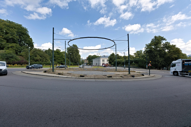 Kreisverkehr in einer Stadt mit Fahrzeugen, Pfählen, Bäumen, Gebäuden und einem bewölkten Himmel.