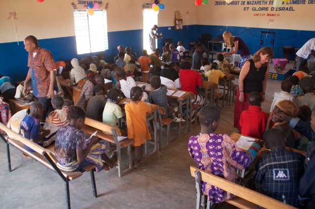 Kinder auf Bänken in der Vordergrund, mit Erwachsenen, die sie führen, und im Hintergrund Stühle, eine Tür, ein Fenster und ein Tisch, sowie Ballons und Text oben.