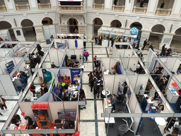 Große Gruppe von Menschen auf einer internationalen Fachmesse mit Ständen, Bannern, Tischen, Stühlen und Luftballons in einem Raum mit architektonischen Bögen, Säulen, Geländern und einem Bildschirm im Hintergrund.
