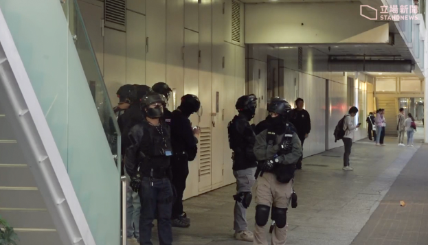 Eine Gruppe von Polizisten in schwarzen Uniformen und Helmen steht vor einem Gebäude, mit einer Pflanze links und ein paar Menschen rechts und verschiedenen Gegenständen im Hintergrund.