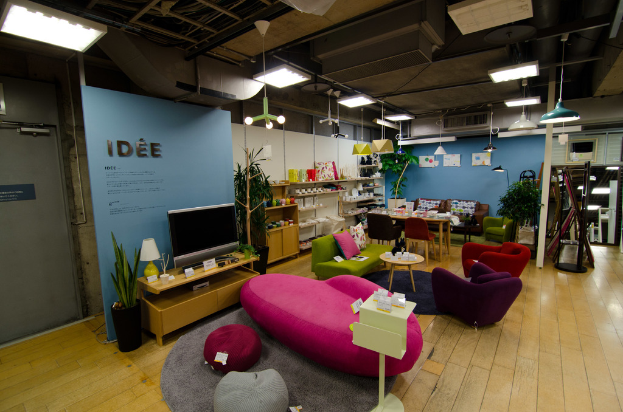 Eine Ladeneinrichtung mit Haushaltsgeräten und Möbeln, darunter ein Fernseher auf einem Tisch, eine rosafarbene Couch, ein Sofa, zwei Stühle um einen kleinen Hocker, ein Esstisch mit Stühlen, ein großes Sofa im Hintergrund und Regalen mit verschiedenen Artikeln, alles beleuchtet von Deckenlampen.