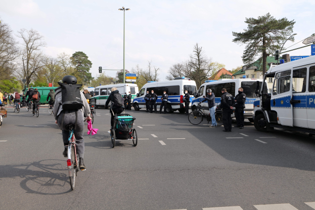 Eine Gruppe von Menschen, die auf Fahrr"adern eine Straäe entlangfahren, die von Polizeiwagen und anderen Fahrzeugen gesäumt ist.