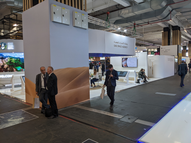 Eine Gruppe von Menschen geht um einen Messestand für die UAE Space Agency, wobei einige Taschen tragen, während im Hintergrund Schilder, Bildschirme und andere Gegenstände sowie Deckenpfeiler und Lampen zu sehen sind.