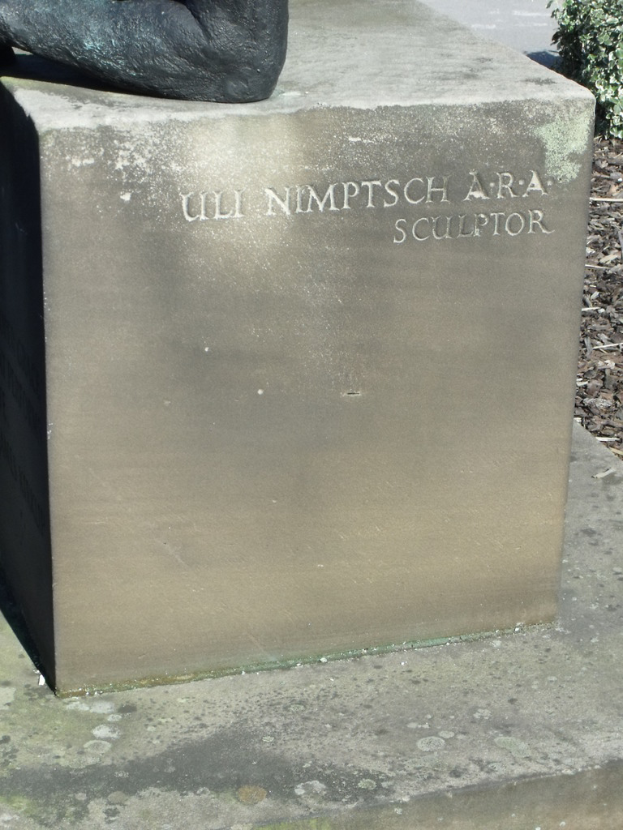 Ein Stein mit Text in Vordergrund, eventuell eine Statue oben, trockene Zweige, Erde und Pflanzen rechts, Straße im Hintergrund.