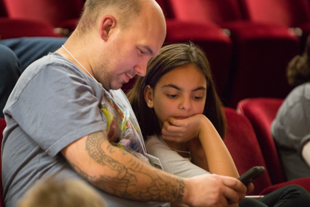 Ein Mann und ein junges Mädchen sitzen in einem Kino, beide konzentrieren sich auf ein Handy, das der Mann hält, mit ein paar anderen Menschen und einem unscharfen Hintergrund.