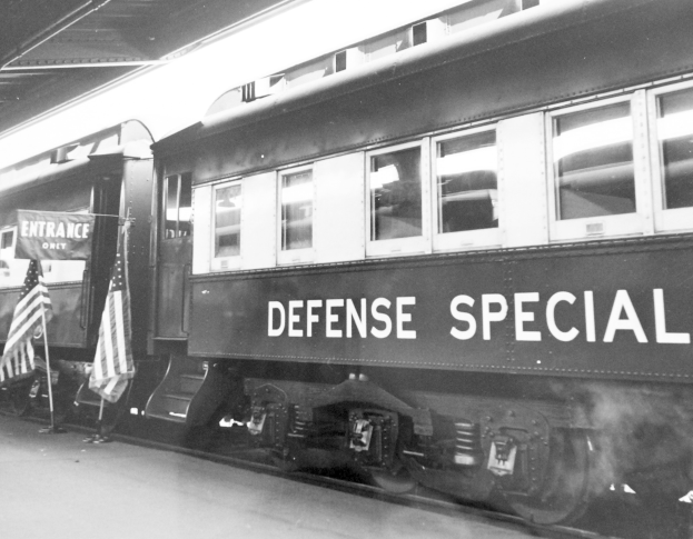 Schwarzes und weißes Foto eines Zuges mit der Aufschrift "Defense Special" auf den Schienen mit zwei Flaggen links und einer Decke oben.