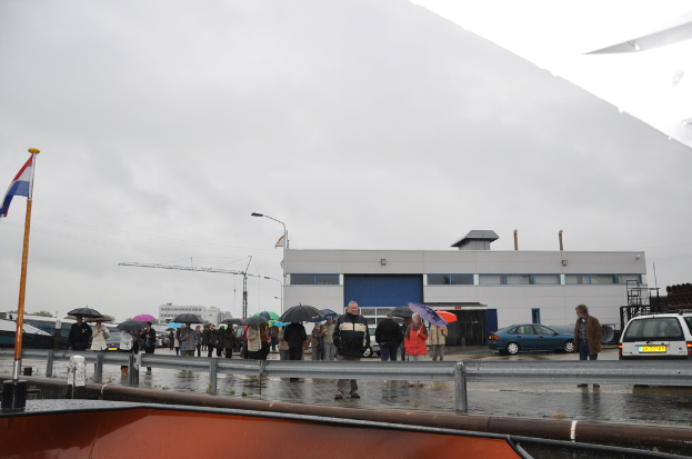 Eine Gruppe von Menschen im Freien, einige mit Schirmen, eine Fahne an einem Mast, zwei Autos, ein Gebäude mit Fenstern, ein Kran, eine Straßenlaterne und eine Absperrung.