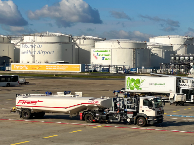 Ein großer weißer und grüner Tanklastzug steht vor einem Gebäude am Frankfurter Flughafen, umgeben von anderen Fahrzeugen, großen weißen Tanks und einem gelben Schild im Hintergrund unter einem klaren blauen Himmel.